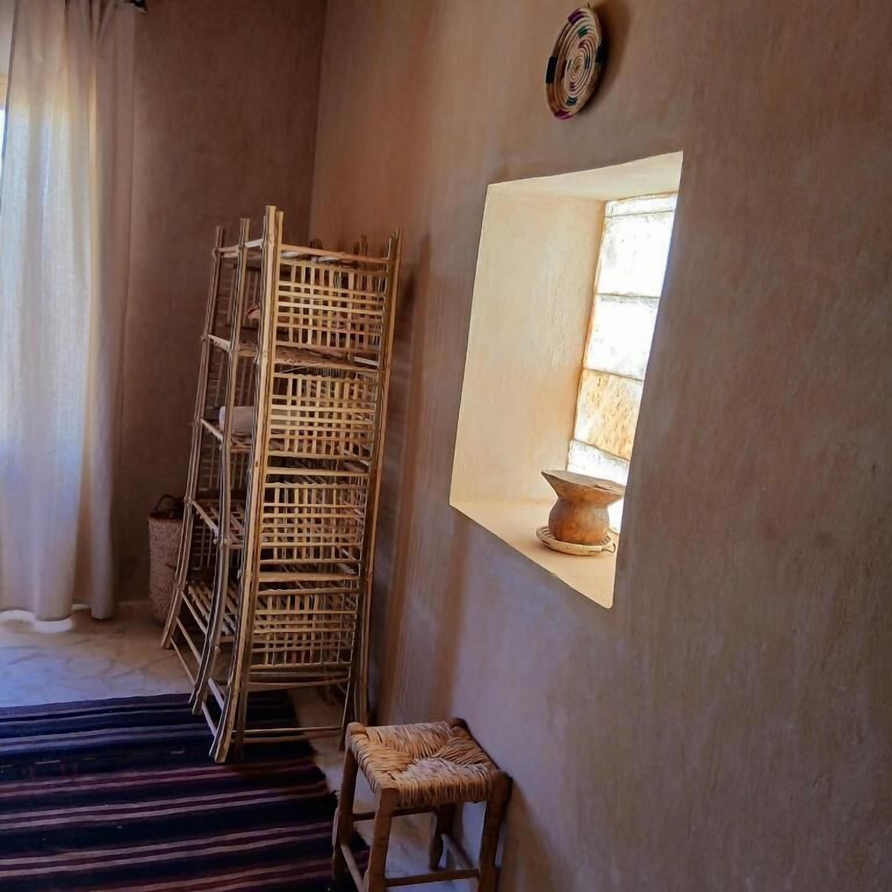 A room with a stack of wicker furniture, a small stool, a wall-mounted shelf with a decorative item, and a window with natural light.