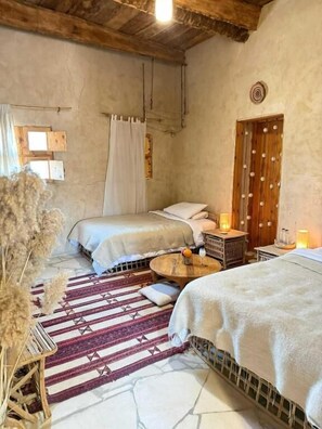 A room with two beds, a round coffee table, a wooden door with circular handles, and a stone floor. - tanirt ecolodge (Siwa)