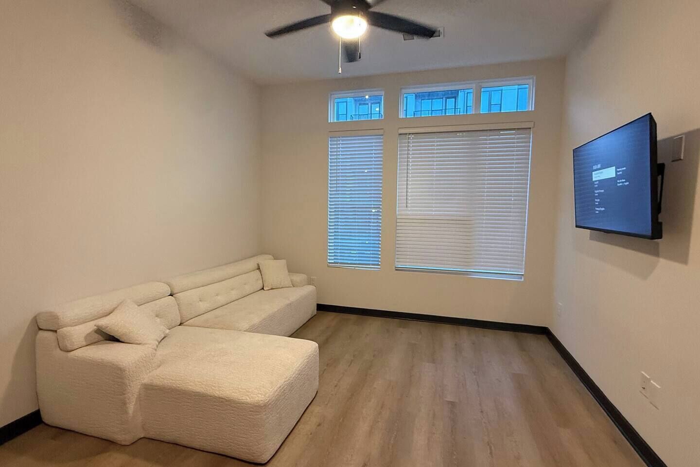 Un salon comprenant un canapé d’angle, un ventilateur de plafond, un téléviseur à écran plat fixé au mur et une fenêtre avec des stores.