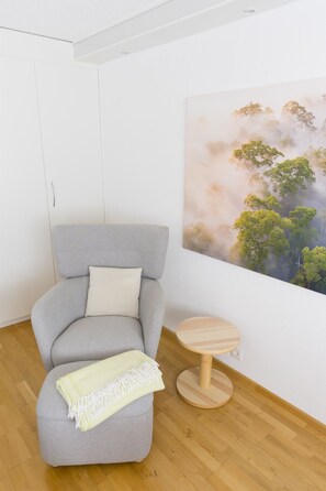 Una habitació amb una butaca gris, una taula rodona de fusta i un gran quadre mural amb arbres.
