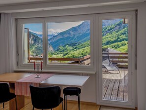 A room with a table and chairs, large windows with a mountain view, and a balcony with a wooden deck. - Olympia 54 # 2-bed apartment, Dusche, WC 2-Bettwohnung, Dusche, WC by Interhome (Leukerbad)