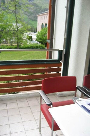 A balcony with a view of a building and greenery. There are red chairs and a white table. - La Promenade 22 # 2-bed apartment, Dusche, WC 2-Bettwohnung, Dusche, WC by Interhome (Leukerbad)
