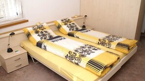 Yhden hengen sänky, jossa on keltainen ja valkoinen kukkapuvukuinen päiväpeite, kaksi yöpöytää, lamppu ja ikkuna, jossa on verhot.