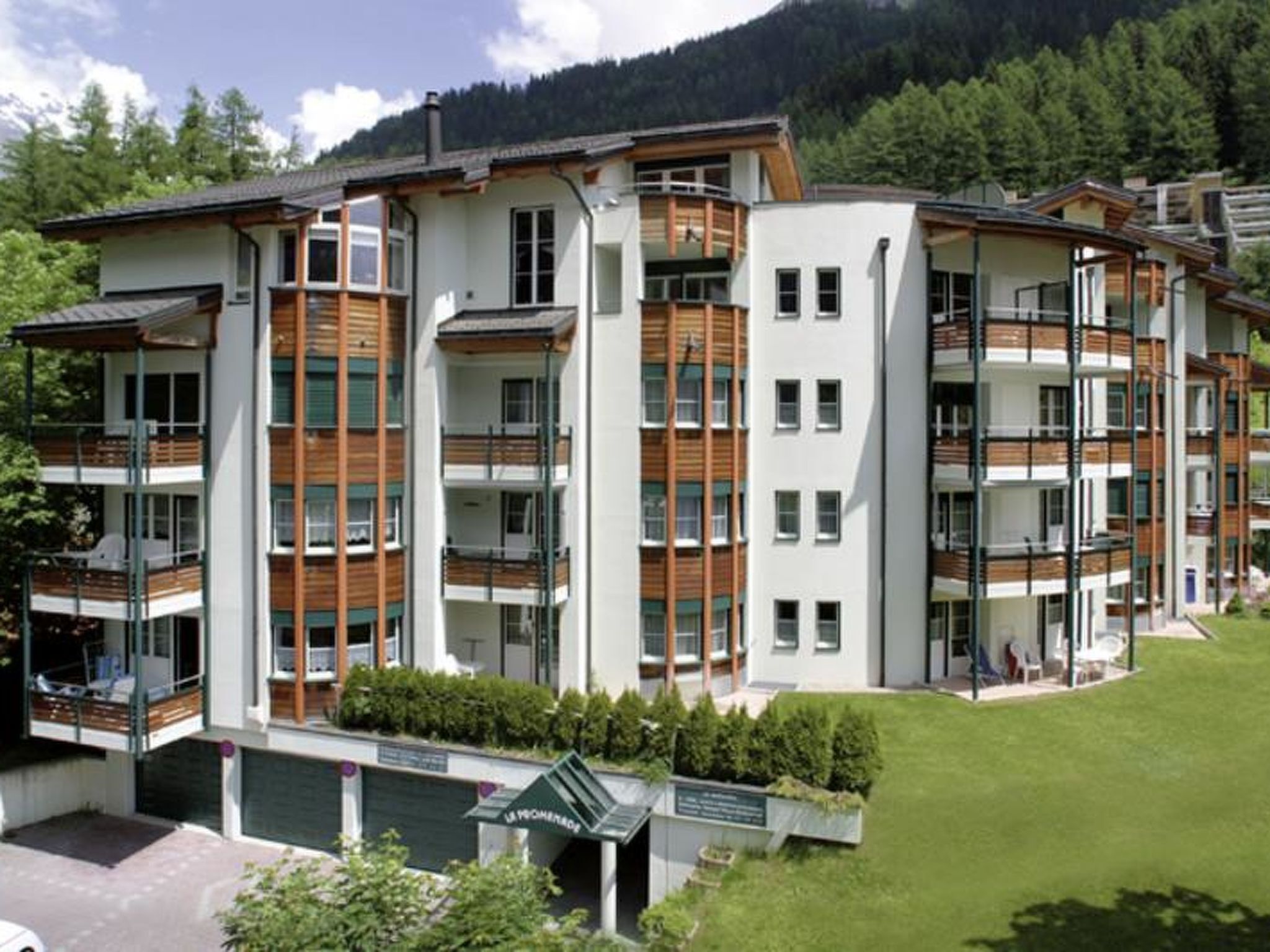 A multi-story residential building with balconies, a green lawn, and a mountain backdrop.