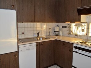 A compact kitchen with wooden cabinets, a white countertop, a built-in dishwasher, a stove, and a coffee maker. - Albina 43 # 4-bed apartment, Dusche, WC 4-Bettwohnung, Dusche, WC by Interhome (Albinen)