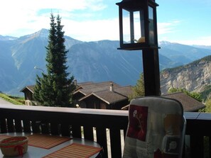 A mountain view from a balcony with a wooden railing, a table with a basket, and a lamp post. - Albina 43 # 4-bed apartment, Dusche, WC 4-Bettwohnung, Dusche, WC by Interhome (Albinen)