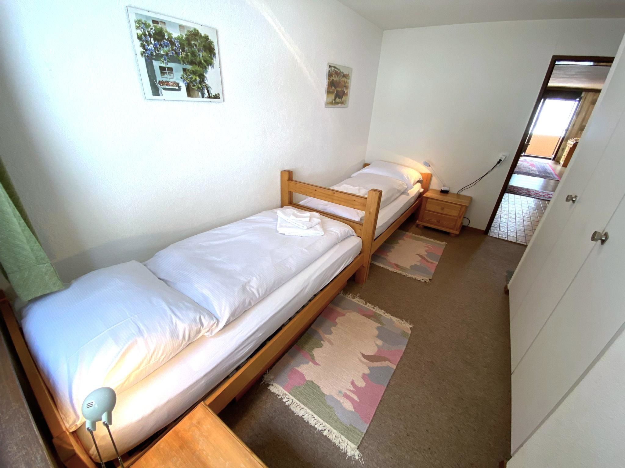 A small, single-room accommodation with two single beds, a bedside table with a lamp, a small rug, and a door leading to a hallway.