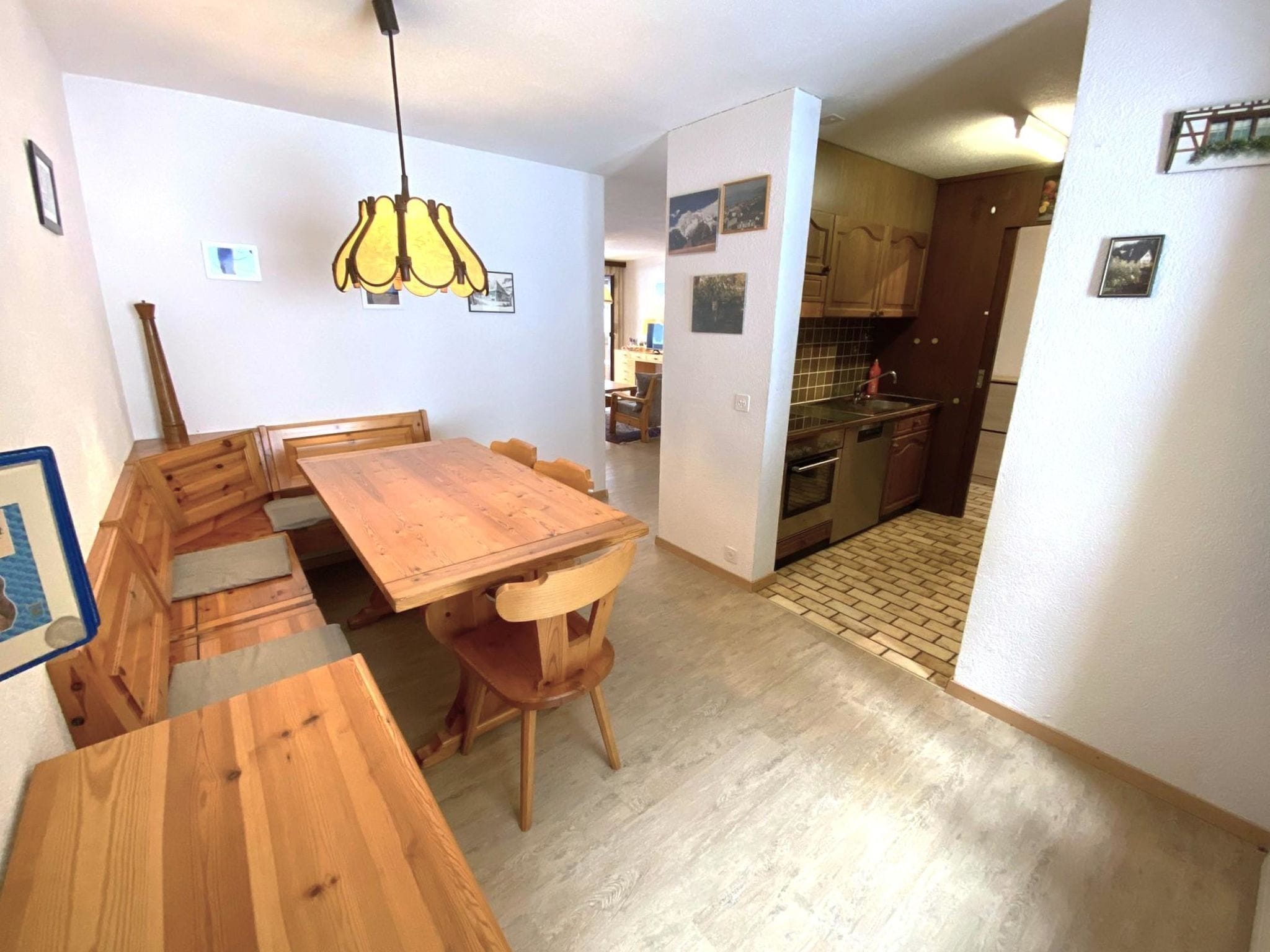 A dining area with a wooden table and chairs, a hanging light fixture, and a kitchen visible through an open doorway.