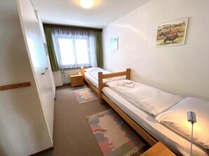 Ein kleines Zimmer mit einem Einzelbett, einem hölzernen Bettgestell, einem Nachttisch mit Lampe, einem Fenster mit Vorhängen und einem an der Wand hängenden Bild.