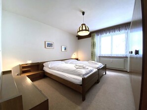A hotel room with a wooden bed, a bedside table, a lamp, a window with curtains, and a picture on the wall. - Lärchenwald 1706 # 6-bed apartment, Dusche, WC 6-Bettwohnung, Dusche, WC by Interhome (Leukerbad)