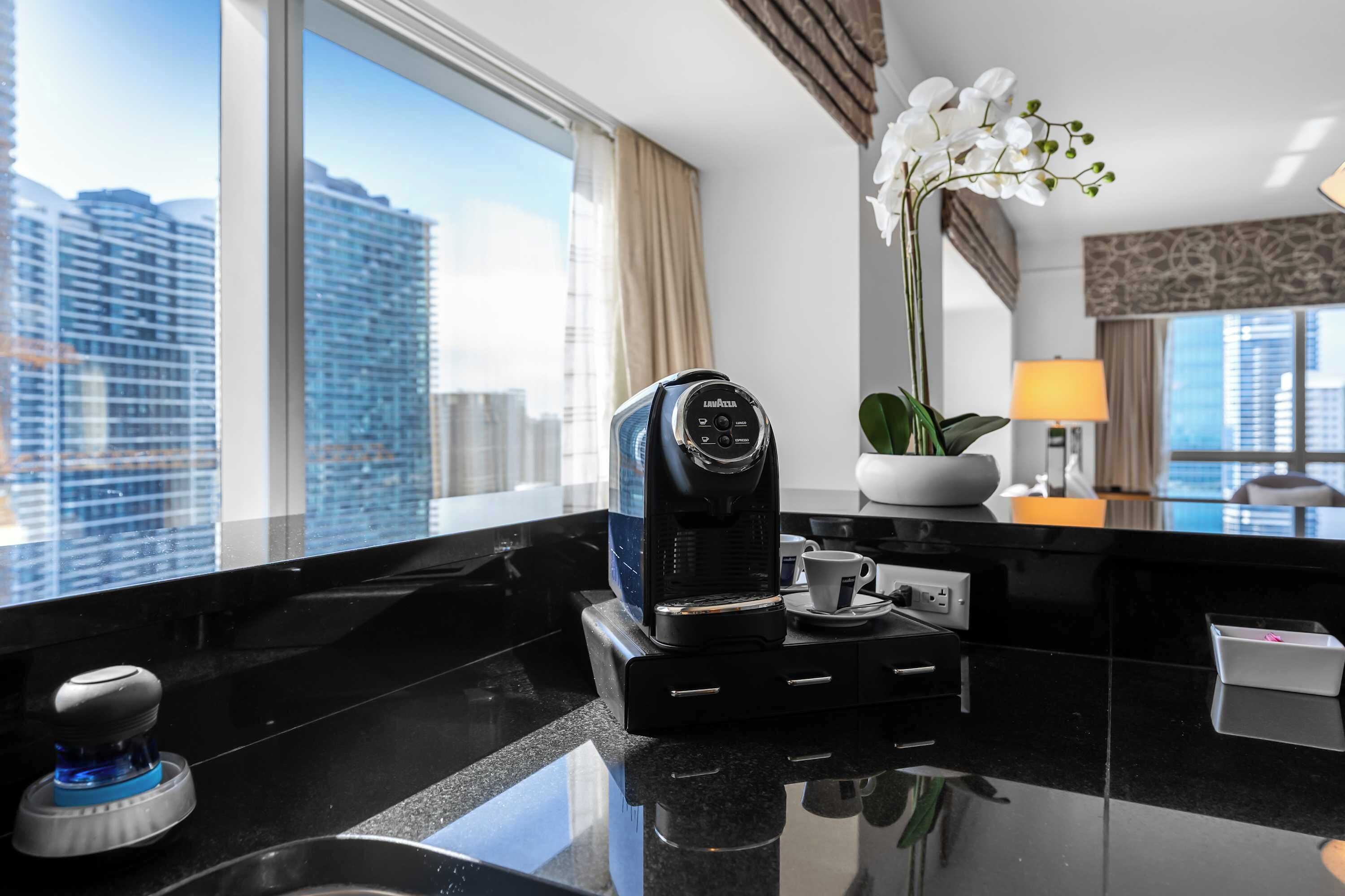 A modern hotel room with a coffee maker, a potted orchid, and a view of the cityscape through large windows.