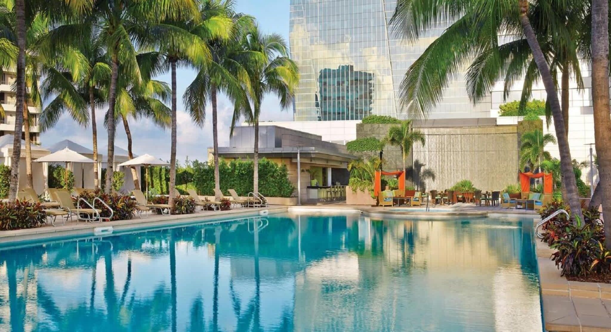 Una piscina con sillones de descanso y sombrillas, rodeada de palmeras y un edificio moderno al fondo.
