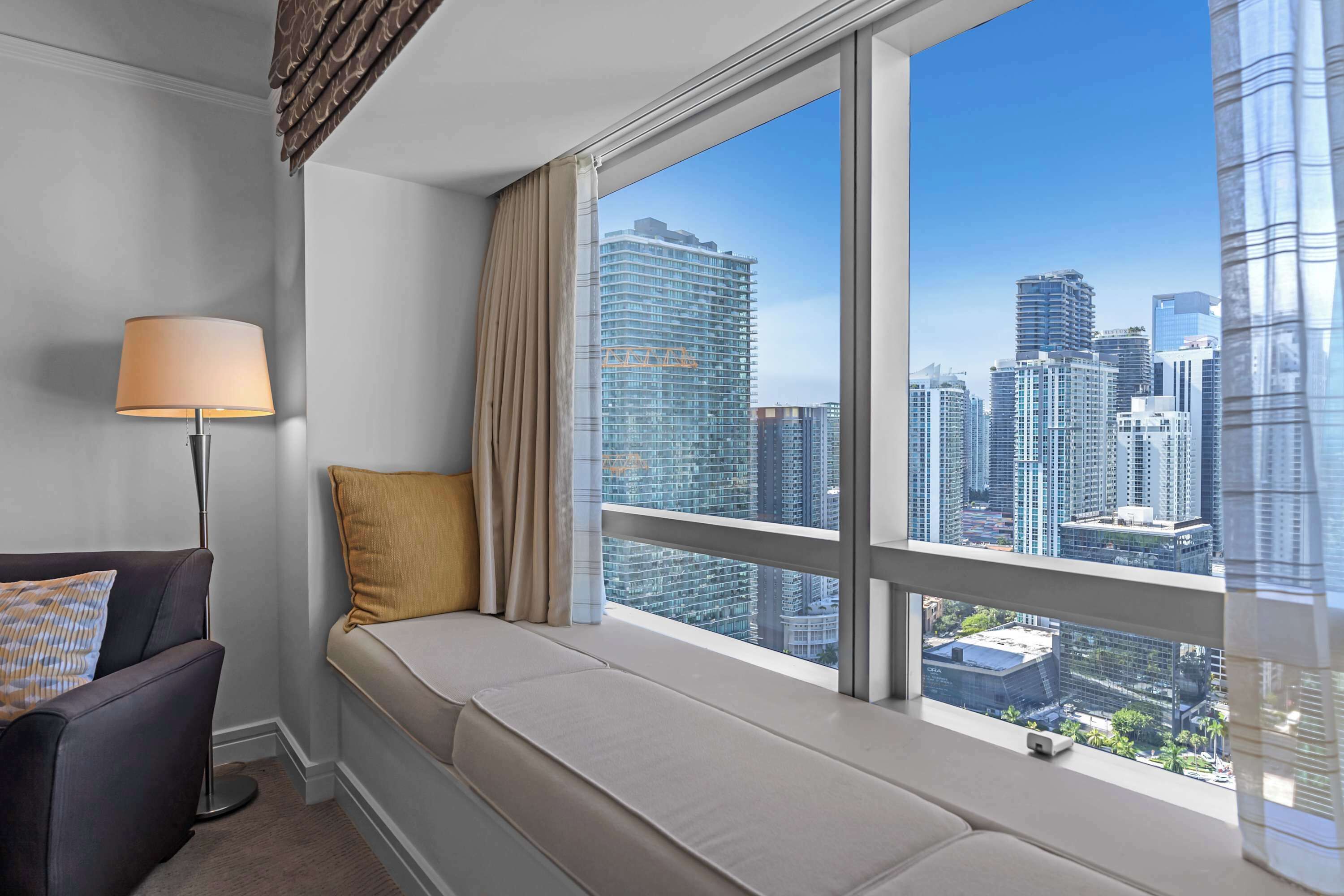 A room with a large window, a sofa, a lamp, and a view of a cityscape.