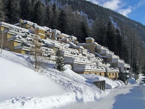 Ein schneebedecktes Gebäude mit mehreren Balkonen, umgeben von bewaldeten Bergen.