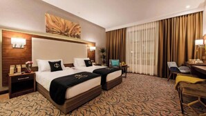 A hotel room with two beds, a desk, a chair, a large window with curtains, and a wall-mounted artwork. - Emerald Twin Delight Sunday Emerald (Jeddah)