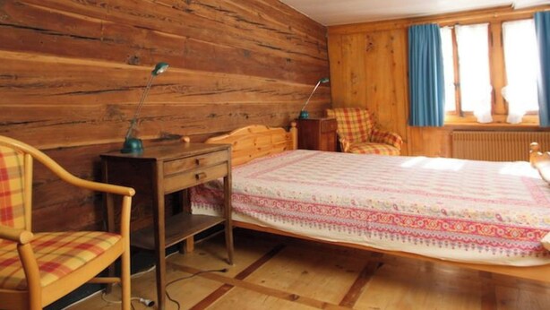 Um quarto com painéis de madeira, uma cama, uma cadeira, uma escrivaninha e uma janela com cortinas.
