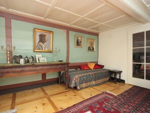 Ein Zimmer mit Holzboden, einem Bett mit gemusterten Betttextilien, einem Tisch mit einem Kandelaber und gerahmten Porträts an der Wand.