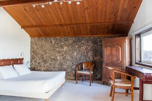 A bedroom with a bed, a wooden chair, a desk, and a stone wall.
