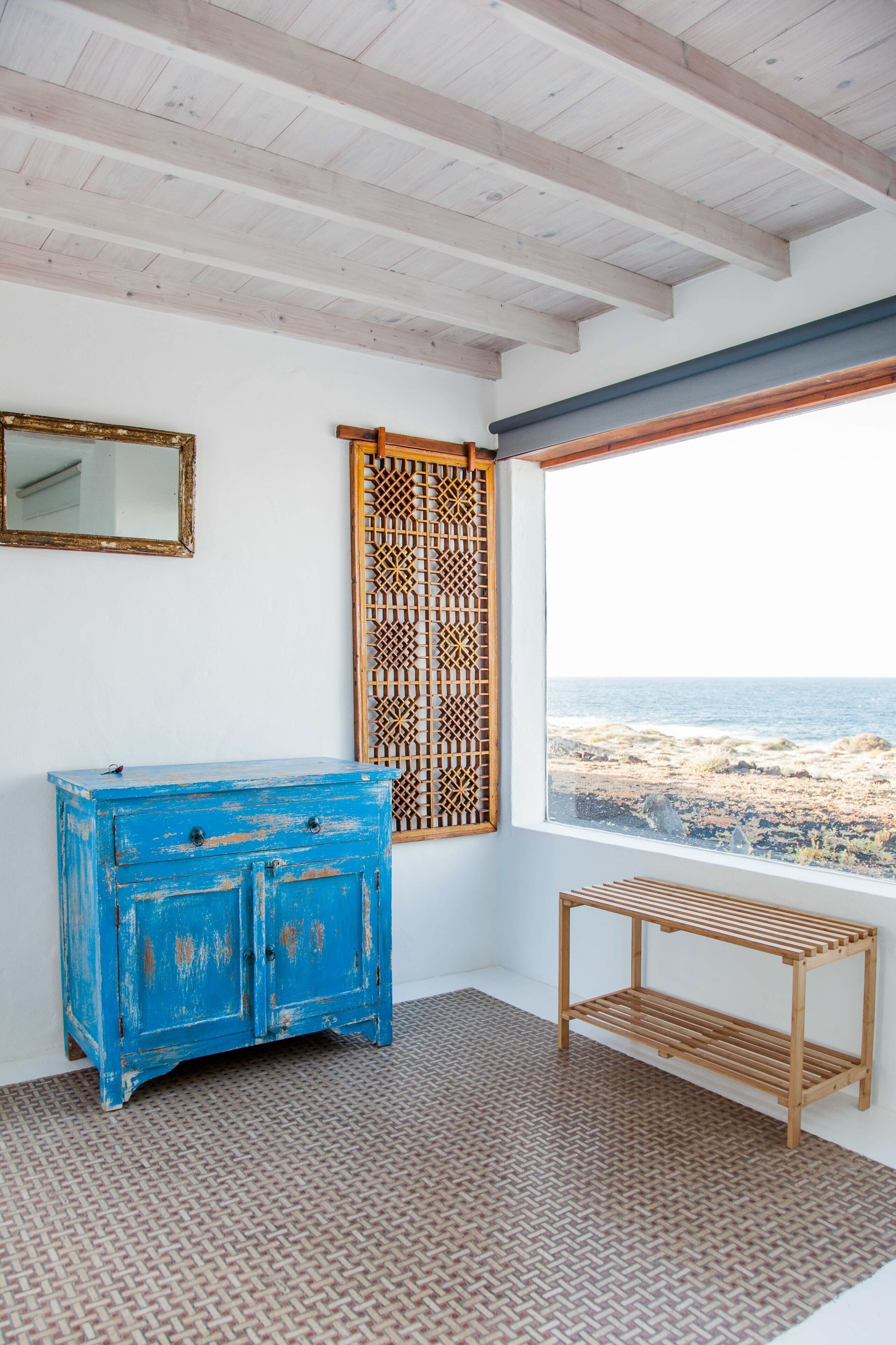 Een kamer met een blauwe kast, een houten plank, een kamerbreed tapijt met een patroon, een raam met uitzicht op het strand en een traliewerk scherm.