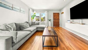 Een moderne woonkamer met een grijze hoekbank, een houten salontafel, een flatscreen televisie en een groot raam met gordijnen.