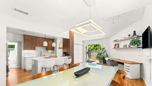 Een moderne keuken met witte kastjes, een eethoek met een glazen tafel, en een aan de muur bevestigde televisie.