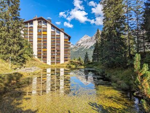 Ein mehrstöckiges Hotel mit Balkonen, vor dem ein See liegt und sich dahinter Berge erheben.