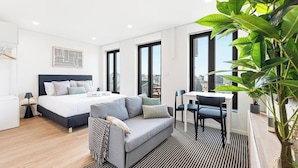 Une chambre d’hôtel moderne équipée d’un lit, d’un canapé, d’une table à manger, de chaises et de grandes fenêtres donnant sur la ville.