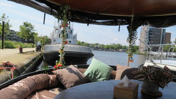 A boat interior with a cushioned seating area, a table with a tissue box, and a view of a canal with a boat named 'WORMERVEE' in the background. - Aloha: An authentic houseboat just 14 minutes from Amsterdam and close to Zaanse Schans (Wormer)