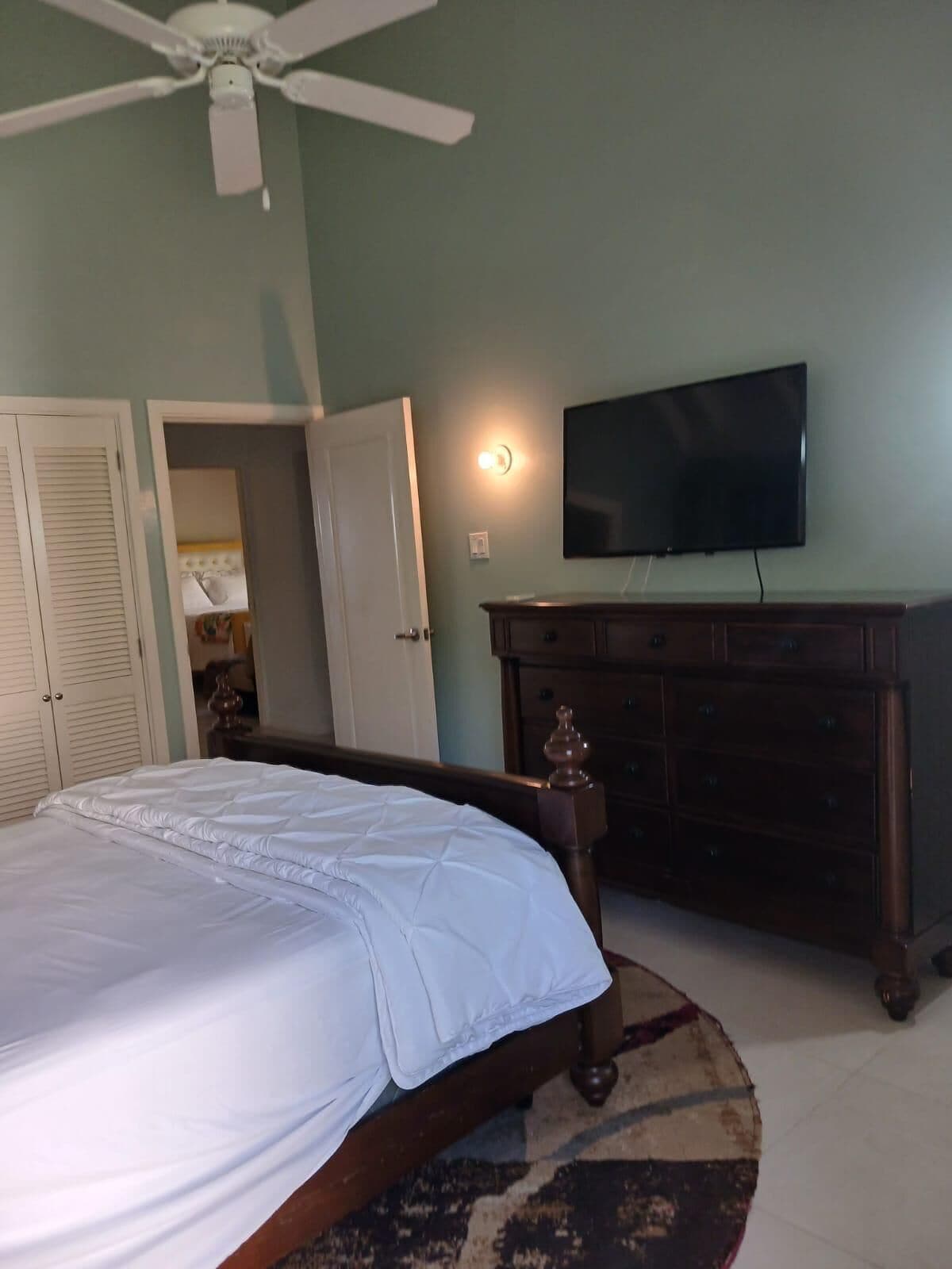 Een slaapkamer met een bed, een plafondventilator, een televisie, een dressoir en een deur naar een andere kamer.