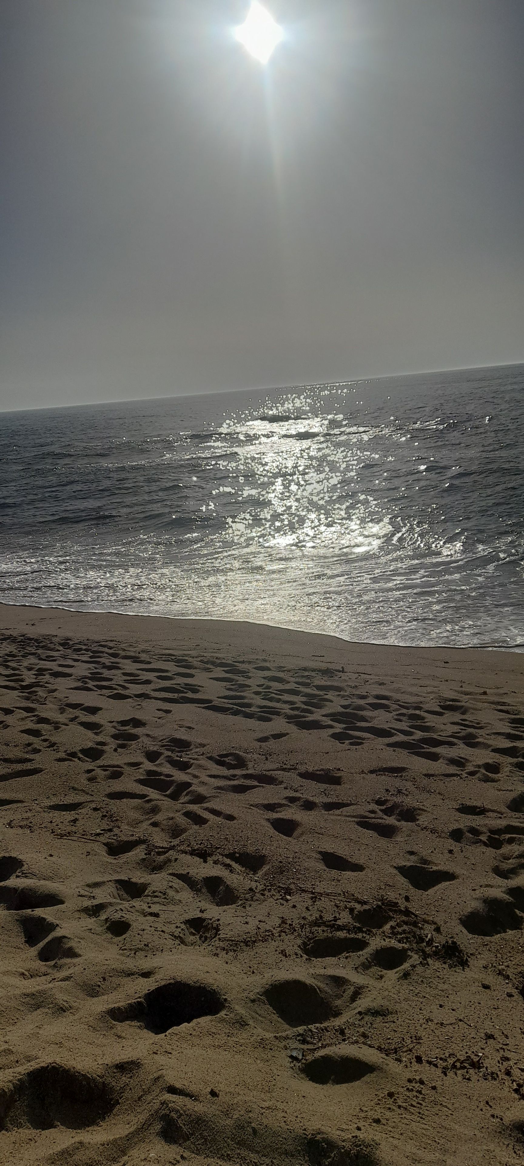 A beach with footprints leading to the water, a clear sky, and the sun shining.