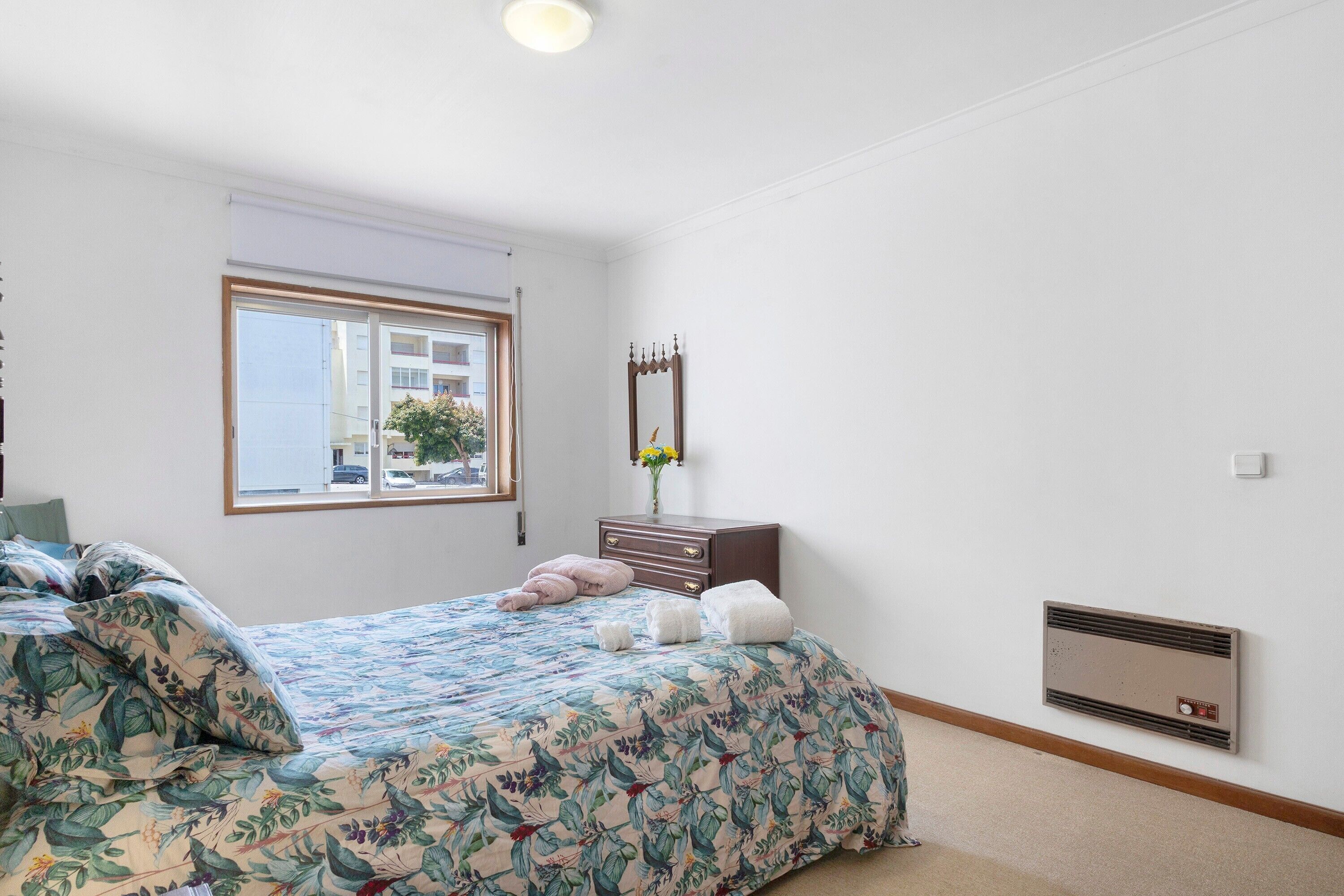 Een slaapkamer met een bed, een ladekast, een spiegel, een raam en een wandheater.
