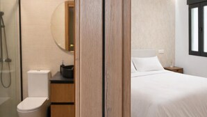 Ein Hotelzimmer mit Holztür, Toilette in Weiß, Waschbecken mit Spiegel, Bett mit weißer Bettwäsche und Fenster mit Jalousien.