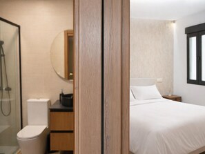 Ein Hotelzimmer mit Holztür, Toilette in Weiß, Waschbecken mit Spiegel, Bett mit weißer Bettwäsche und Fenster mit Jalousien.