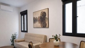 Ein Wohnzimmer mit einer Couch, einem runden Tisch und zwei Stühlen. An der Wand hängt ein Gemälde und ein Fenster mit Jalousien.