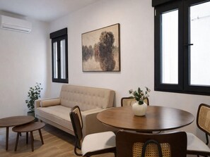 Ein Wohnzimmer mit einer Couch, einem runden Tisch und zwei Stühlen. An der Wand hängt ein Gemälde und ein Fenster mit Jalousien.