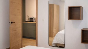 Ein Schlafzimmer mit einem Bett, einer Holztür, einem Spiegel, einer kleinen Holzablage und einer Küchenzeile im Hintergrund.