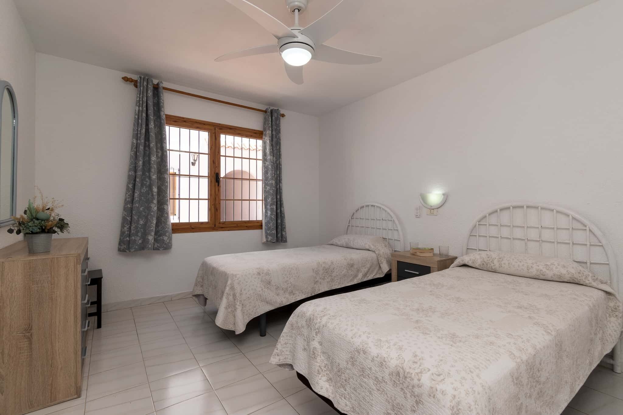 A room with two beds, a ceiling fan, a window with curtains, a bedside table, and a dresser.