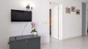 A hallway with a flat-screen TV mounted on the wall, a grey cabinet with a decorative emblem, a potted pink orchid, and three framed pictures on the wall. - Port Royale 75 - Two Bed (Los Cristianos)