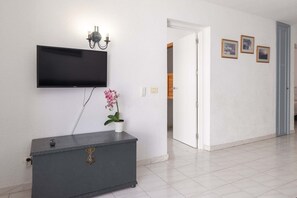 A hallway with a flat-screen TV mounted on the wall, a grey cabinet with a decorative emblem, a potted pink orchid, and three framed pictures on the wall. - Port Royale 75 - Two Bed (Los Cristianos)