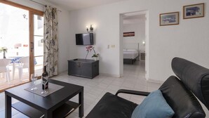 A living room with a sofa, a table with a bottle and glass, a TV, a bed, and a door leading to another room. - Port Royale 75 - Two Bed (Los Cristianos)