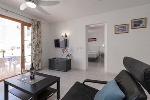 A living room with a sofa, a table with a bottle and glass, a TV, a bed, and a door leading to another room. - Port Royale 75 - Two Bed (Los Cristianos)