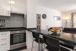 A modern kitchen with a built-in oven, a dining table with chairs, a fruit bowl, a clock, and a bookshelf. - Port Royale 75 - Two Bed (Los Cristianos)