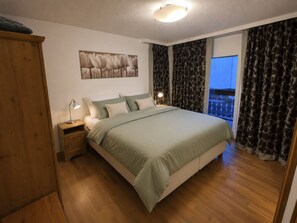 A bedroom with a bed, a nightstand, a lamp, a wardrobe, a painting on the wall, and a view of the outside through the window. - Ambassador B33 # 4-bed apartment, Dusche und Bad, WC by Interhome (Leukerbad)