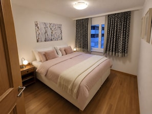Quarto com cama, mesas de cabeceira, luminária de teto e cortinas na janela.