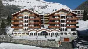 Un hotel de varios pisos con balcones, fachada de piedra y montañas al fondo.
