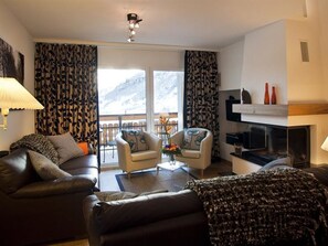 Ein Wohnzimmer mit einer Couch, einem Sessel und einem Couchtisch. Ein Kamin und ein Fernseher sind ebenfalls vorhanden.