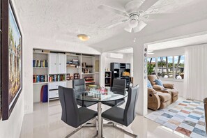 Un salon moderne avec une table à manger en verre, des chaises noires, un ventilateur de plafond et une bibliothèque remplie de livres et d’objets décoratifs.