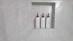 Een badkamer met een marmeren vloer en een plank met drie flessen, gelabeld 'Body Wash', 'Shampoo' en 'Conditioner'.