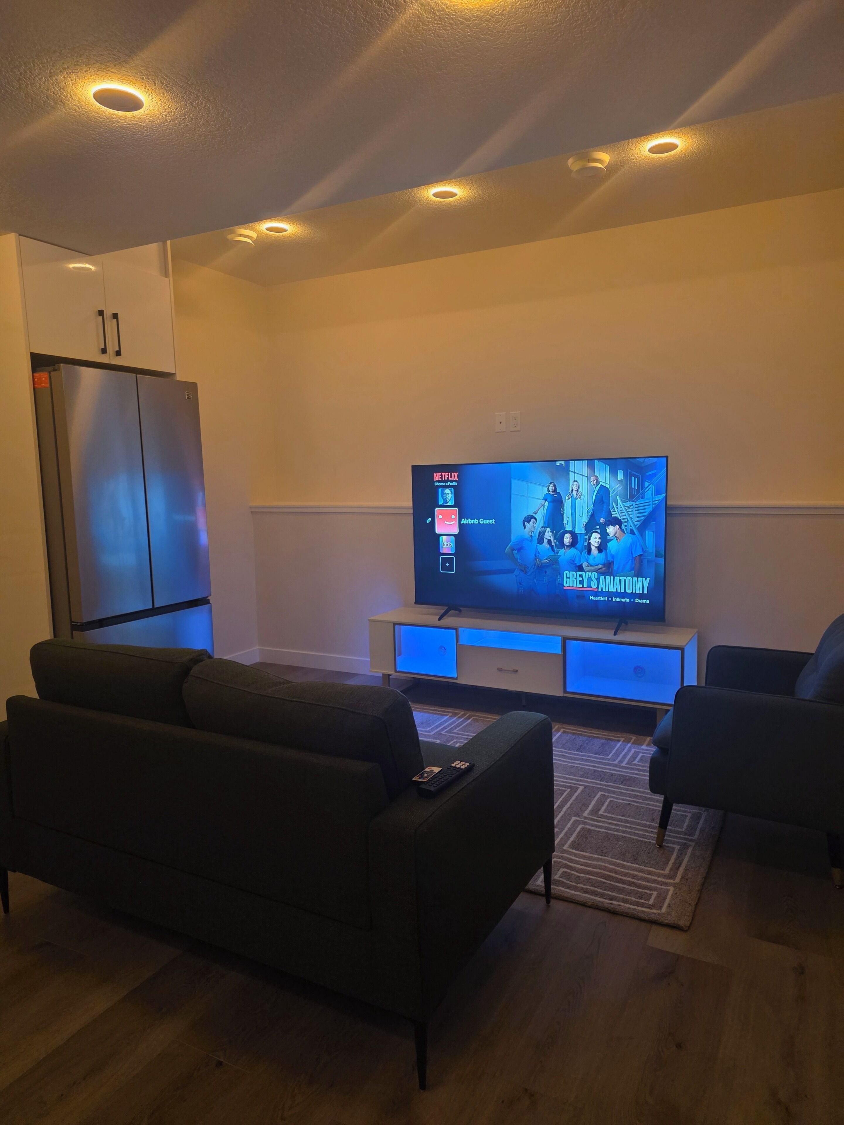 A living room with a flat-screen TV, two sofas, a wooden floor, and a built-in refrigerator.