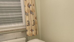 Una habitación pequeña con cama individual, un radiador, una ventana con persianas y una cortina estampada.